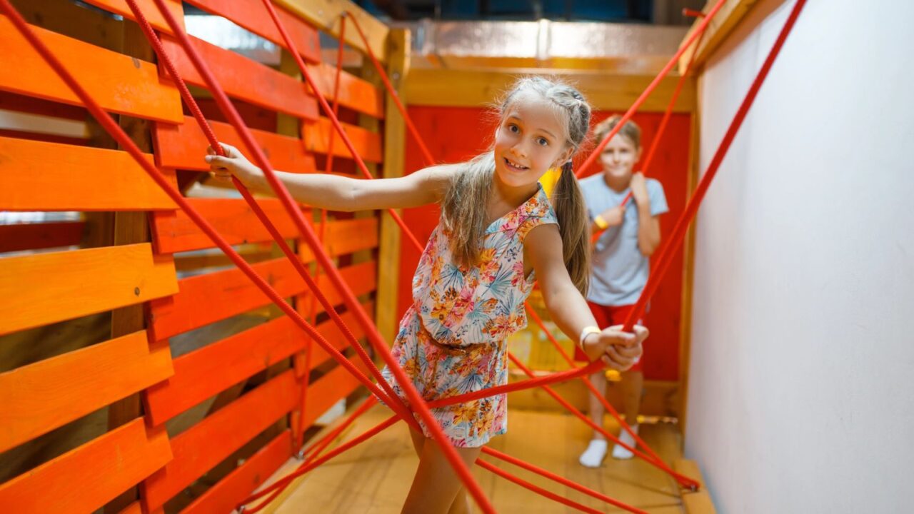 Girl climbing a red safety net structure with a boy following behind in a play area.