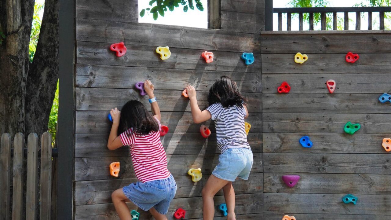 Two children climbing on a wooden wall with colorful handholds outdoors on a sunny day.