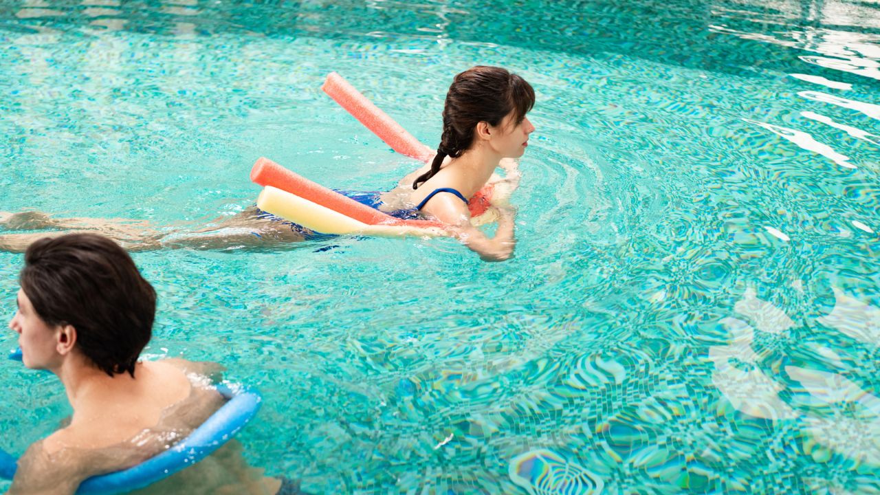 Two women swimming in a pool with pool noodles.