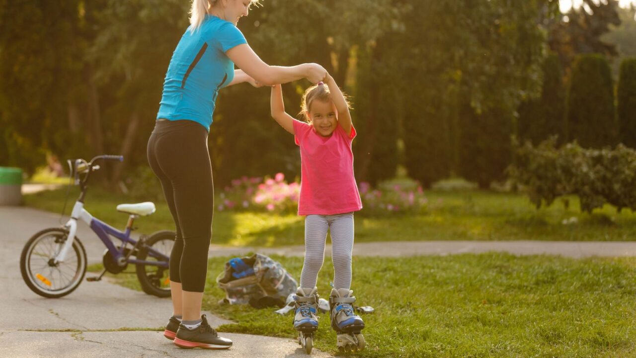 A young girl in a blue shirt helping a smiling girl in a pink shirt to skate outdoors in a park at sunset.