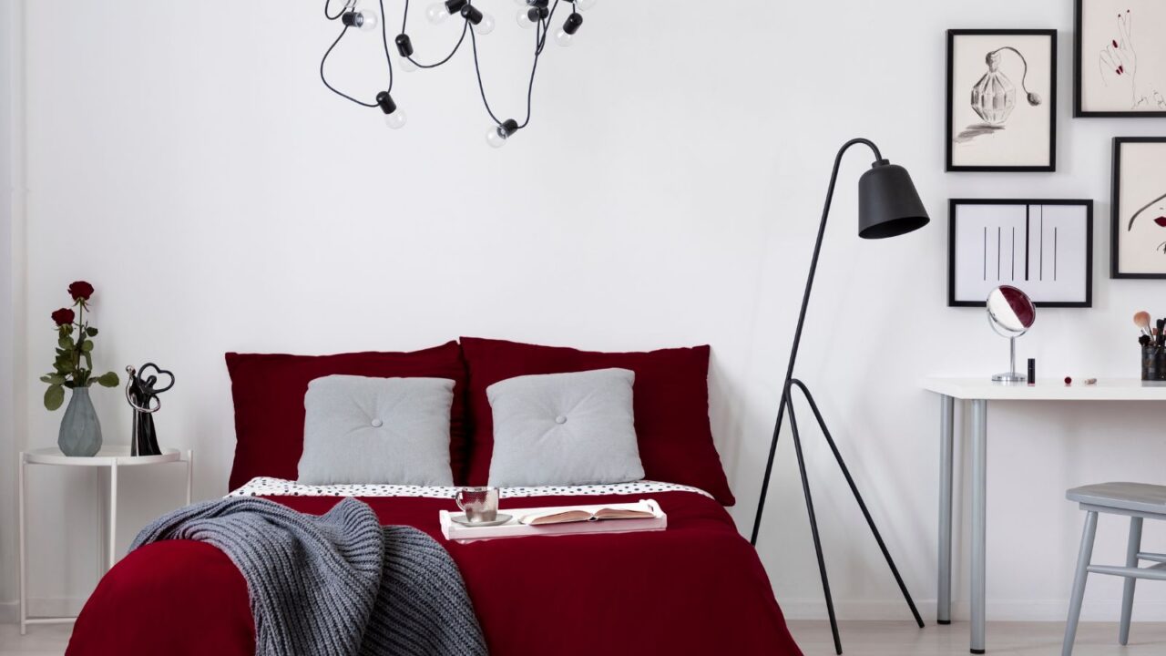 A neutral bedroom interior with burgundy bedding, floor lamp, and side table with flower vase and sculpture.
