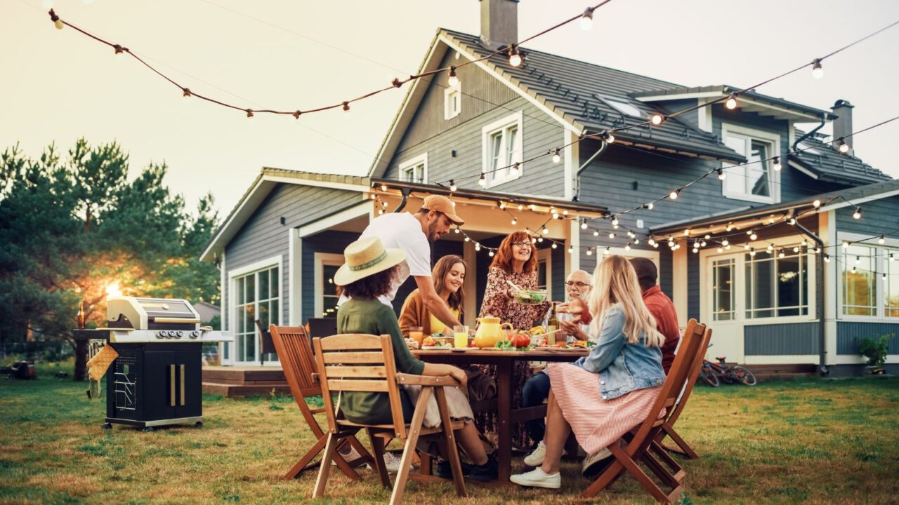 A family enjoying a barbeque party in a garden with illuminated string lights, a grill, and wooden dining setup.