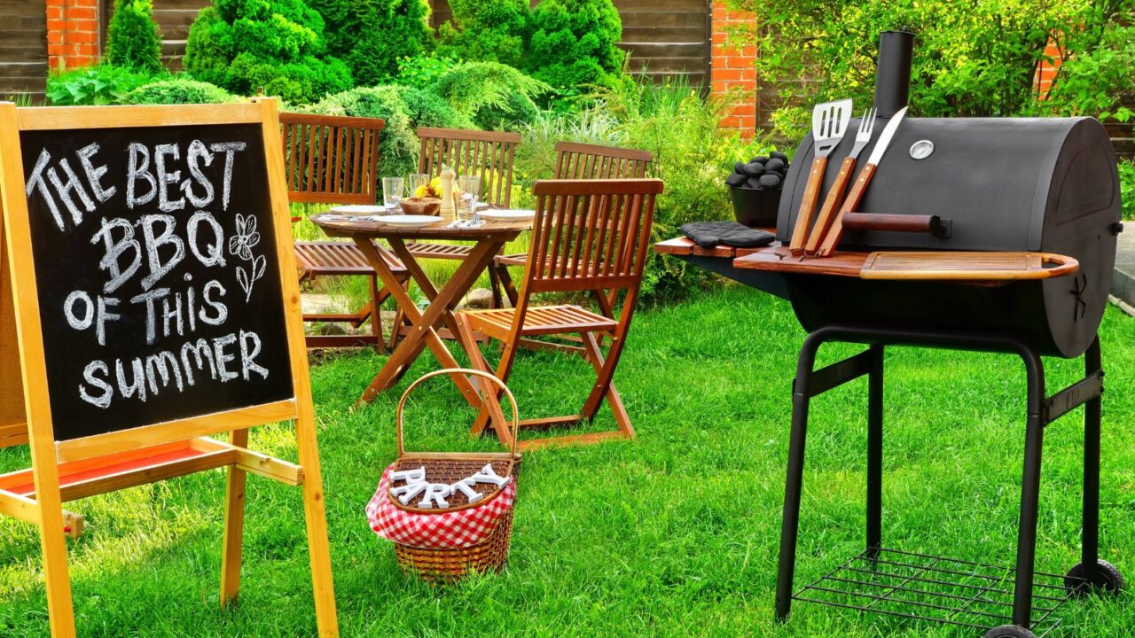 A beautiful outdoor barbeque party setup with wooden chalkboard, grill, wooden dining set, and rustic woven basket on the grass.