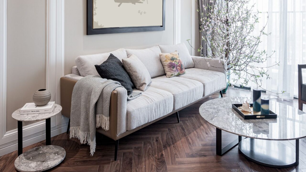 Modern living room with a light gray sofa, marble tables, floral accents, and a tall flowering branch by sheer window curtains.