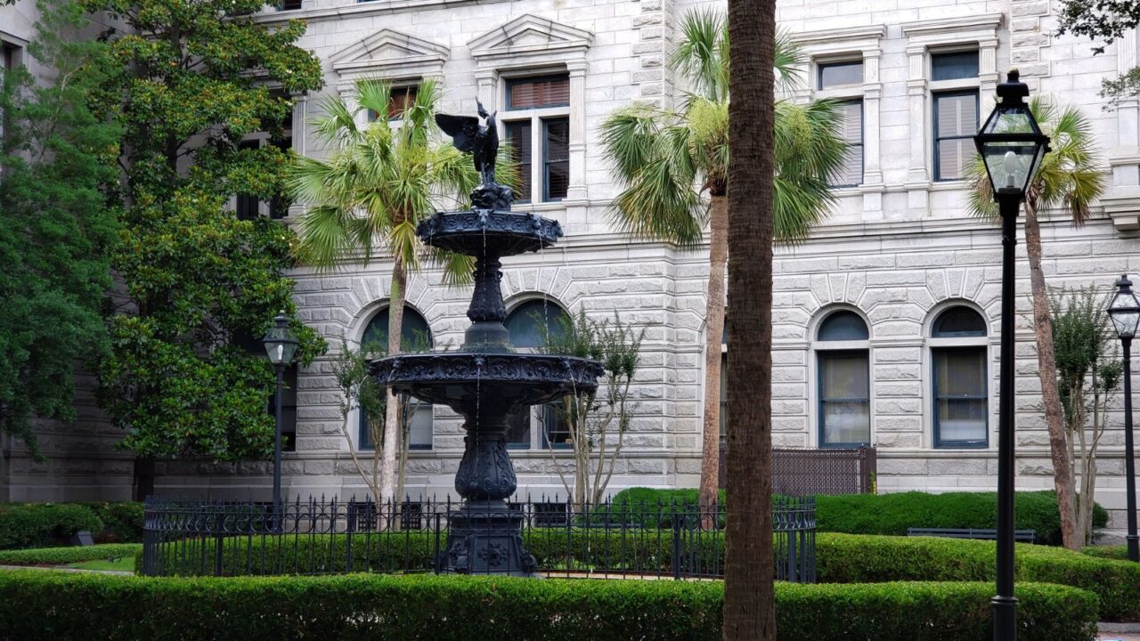 A black, ornate fountain featuring a bird statue atop its highest tier, in front of a classical stone building with arched windows.