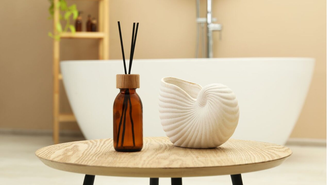 A reed diffuser and a white decorative shell shaped ceramic are placed on a round wooden table in a bathroom setting, with a white bathtub visible in the background.