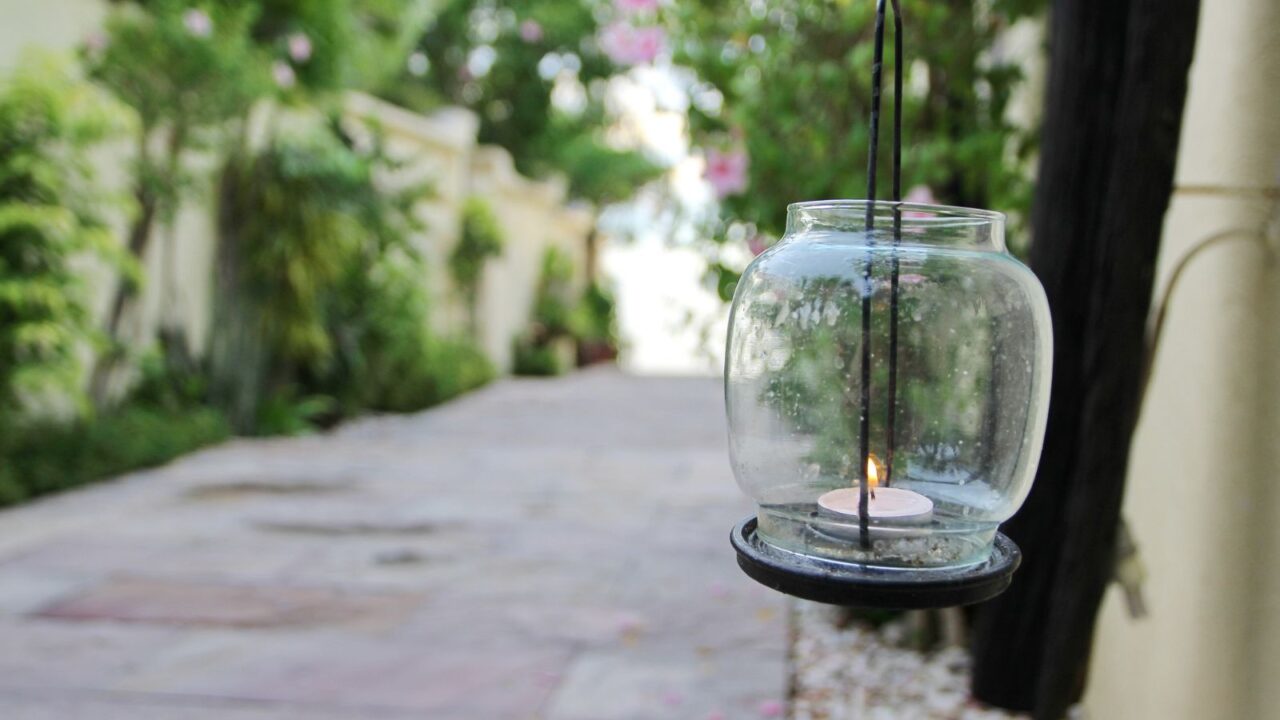 A glass hanging lantern with a small lit candle inside, suspended by black metal wires, set against a blurred garden pathway with greenery and pink flowers in the background.
