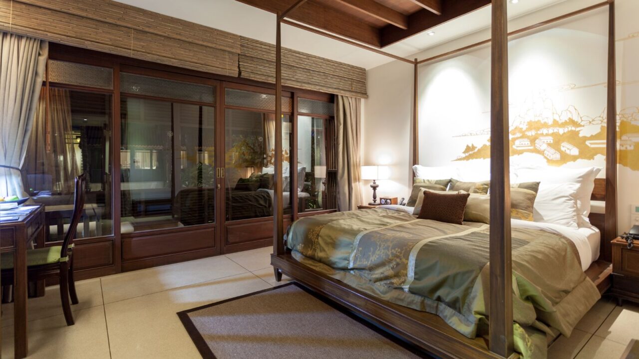 A luxurious bedroom with a four-poster bed, olive green bedding, and a large windowed wall revealing an outdoor area in the Koh Samui resorts in Thailand.
