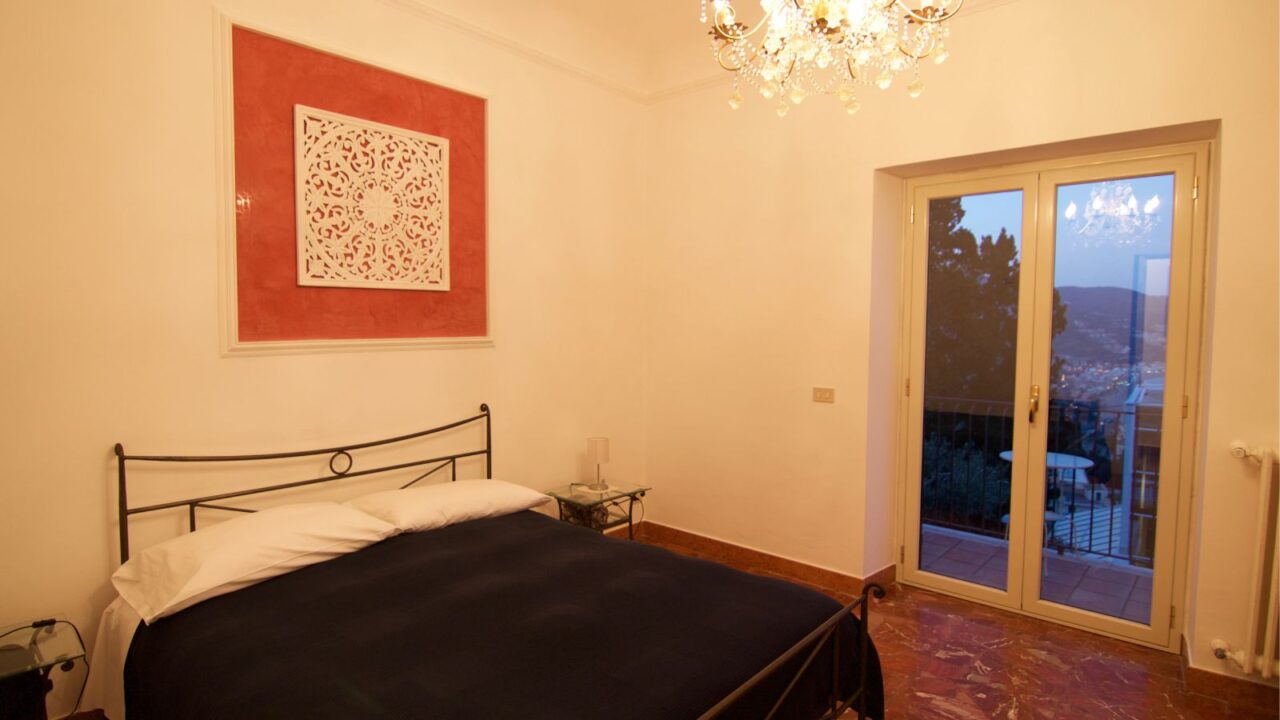 A bedroom with a dark bed, a red and white decorative piece on the wall, a crystal chandelier, and a balcony with a view of hills.