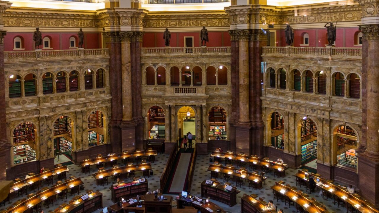 The ornate main reading room of the Library of Congress, featuring rows of desks with lamps, bookshelves lining the walls, and a grand architectural design.
