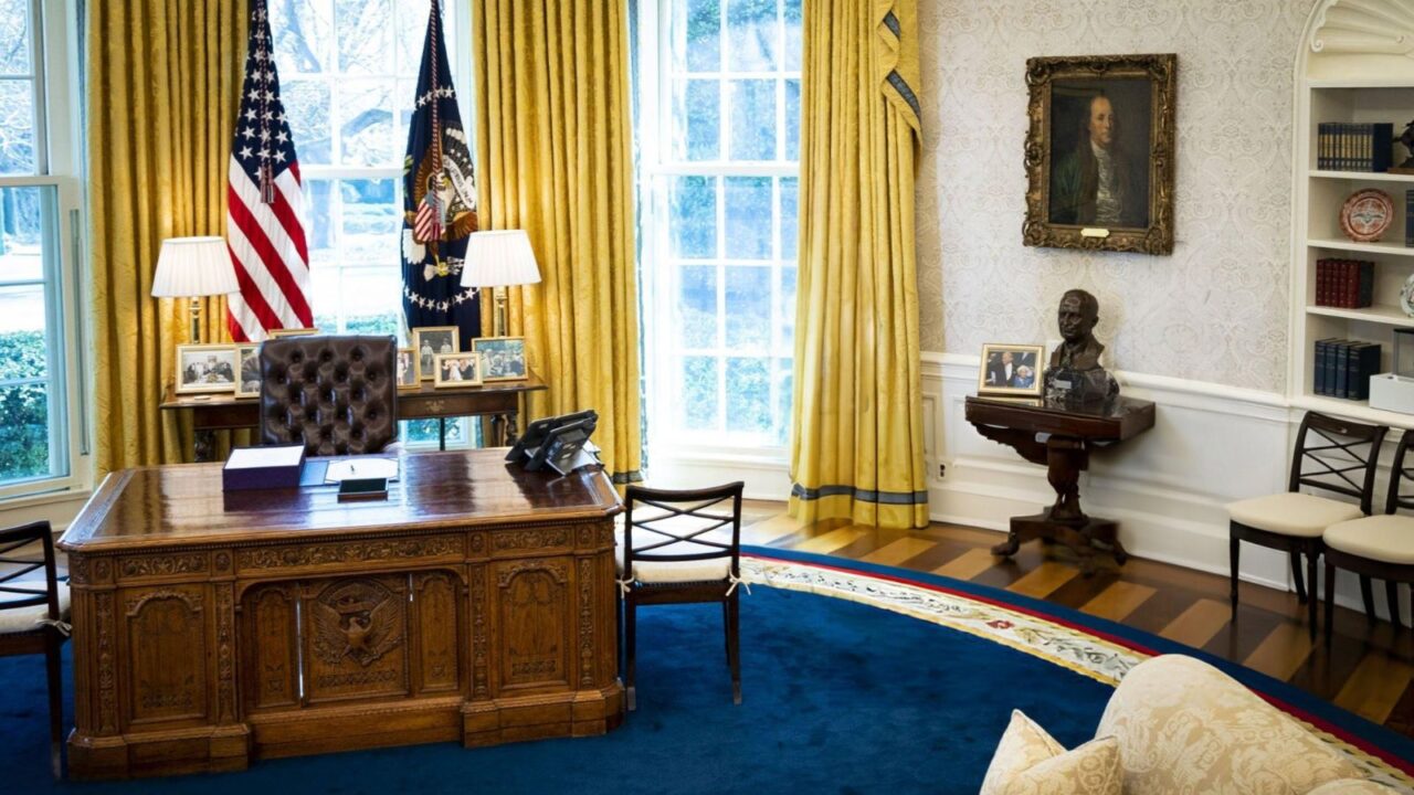 The Oval Office in the White House, featuring the Resolute Desk in the foreground, flanked by the American flag and the Presidential Seal flag, with yellow curtains framing large windows, a portrait on the wall, and a bust on a pedestal.