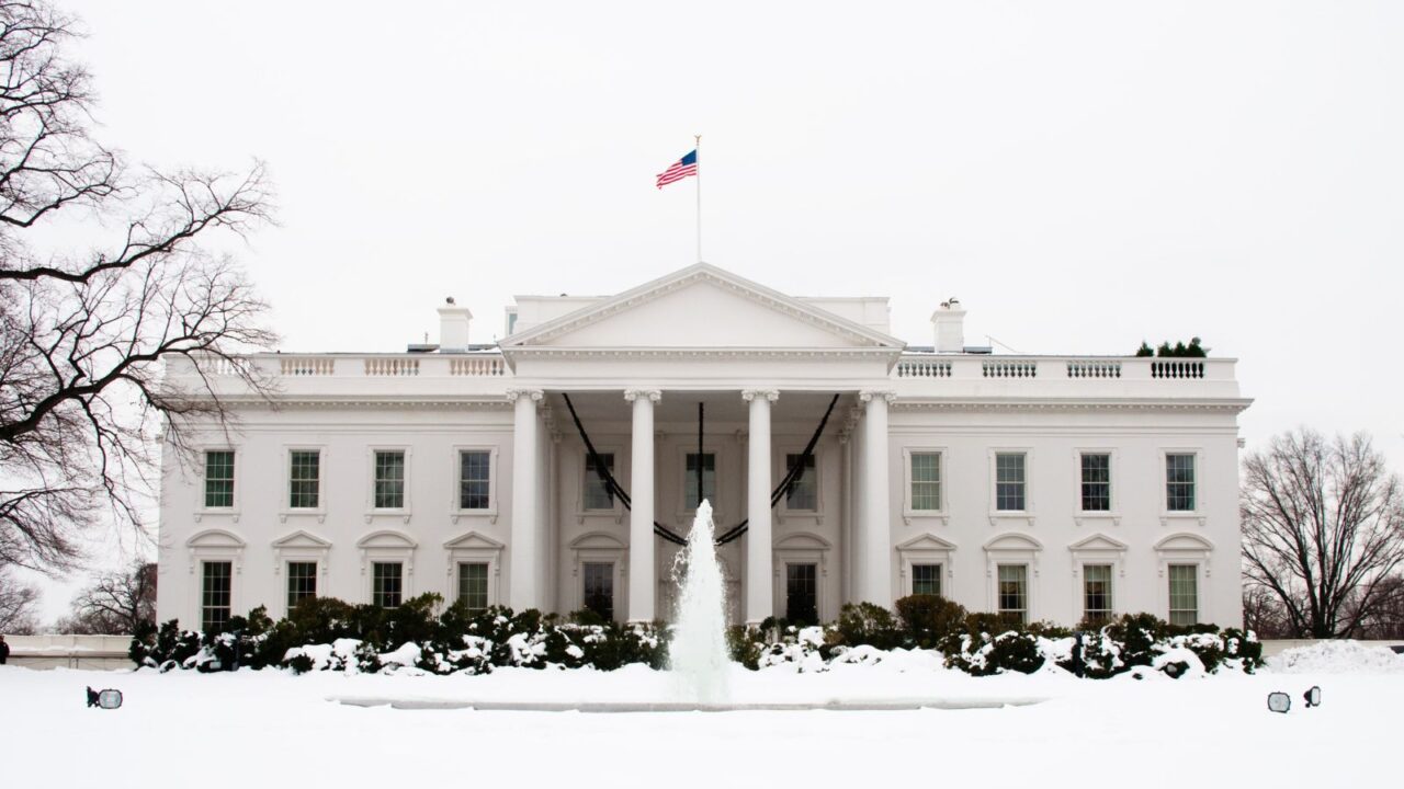 The front view of the White House on a snowy day, with a U.S. flag flying above the roof, a central fountain spraying water, and trees and bushes covered in snow surrounding the building.