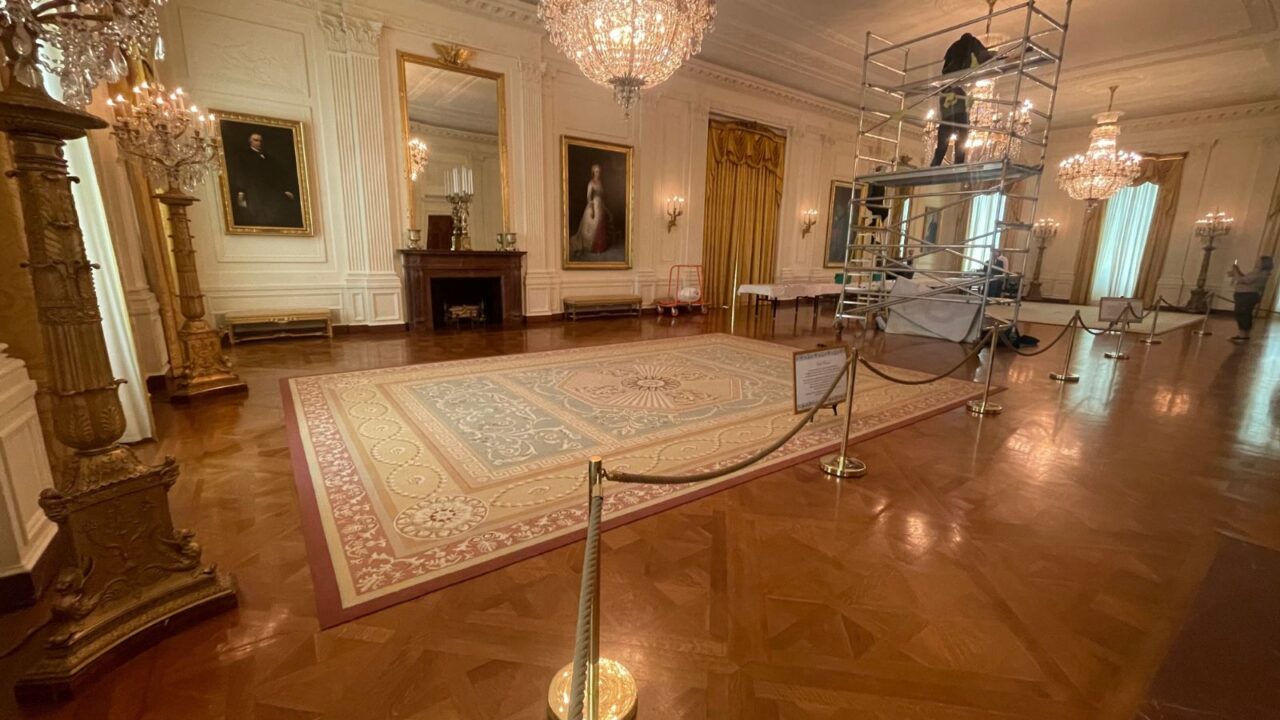 A grand, ornate room, likely in the White House, features chandeliers, large paintings, a fireplace, and a patterned rug, with scaffolding and a worker visible on the right side.