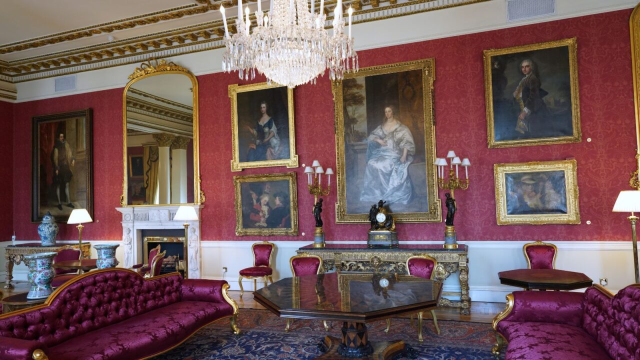 A grand, richly decorated room featuring red patterned wallpaper, multiple large framed paintings, furniture including a long sofa and chairs, and a large chandelier illuminating the space.