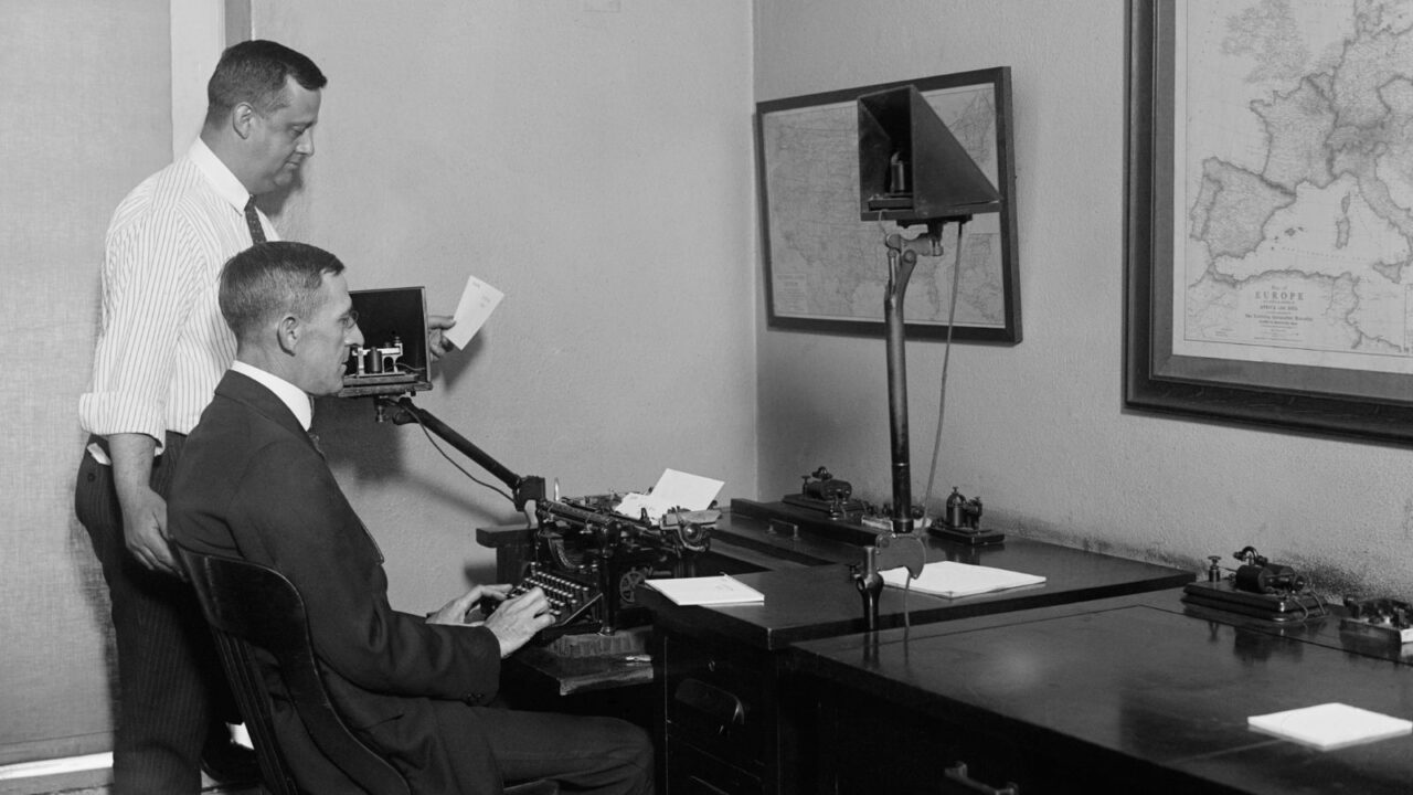 Two men in White House telegraph room, with one man seated at a typewriter and the other standing beside him, surrounded by telegraph equipment and a map.