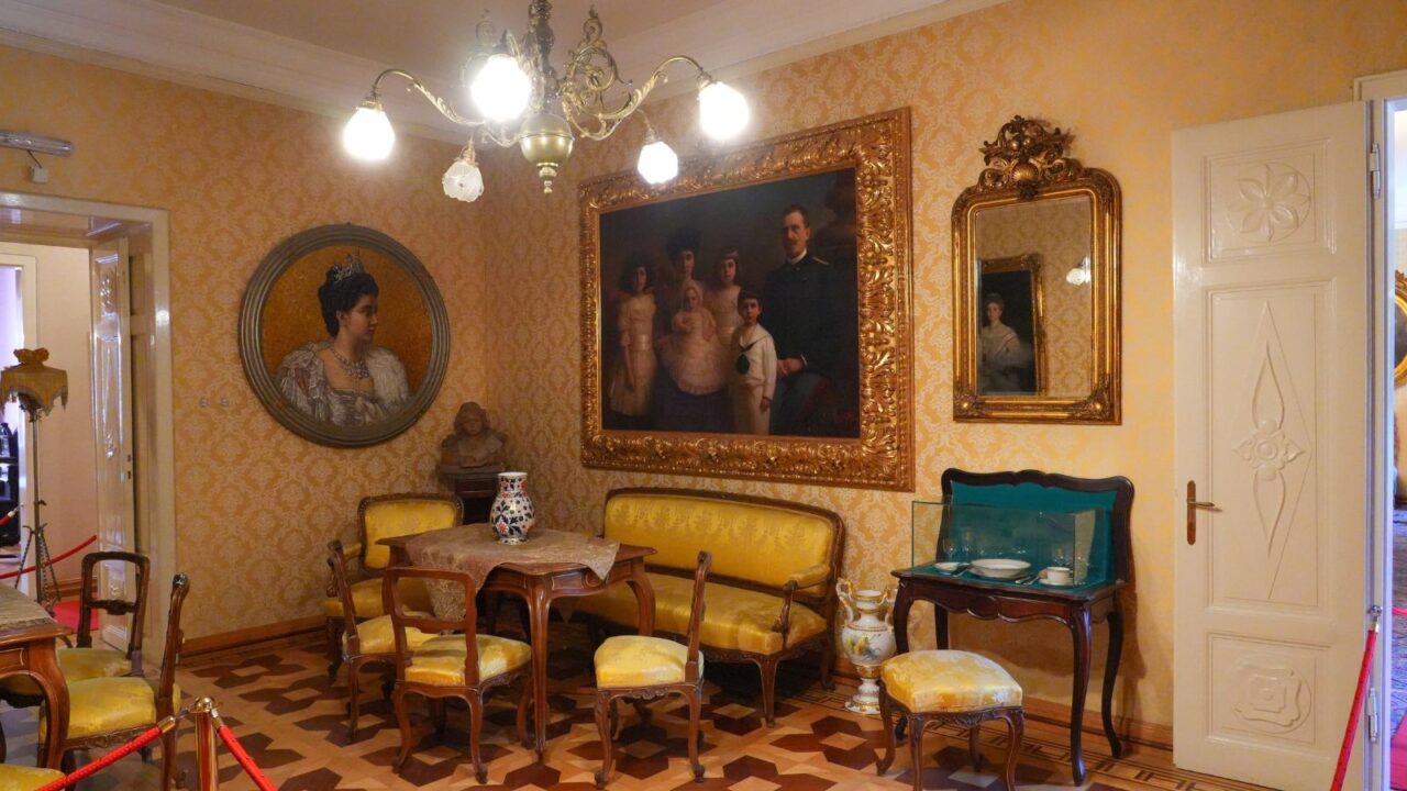 An ornate room, featuring period furniture, framed paintings, and a chandelier.