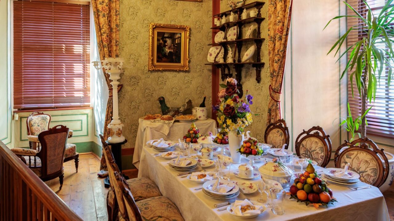 A formal dining room set with a long table laden with dishes and fruit arrangements, featuring ornate furniture, patterned wallpaper, and a large window with wooden blinds.