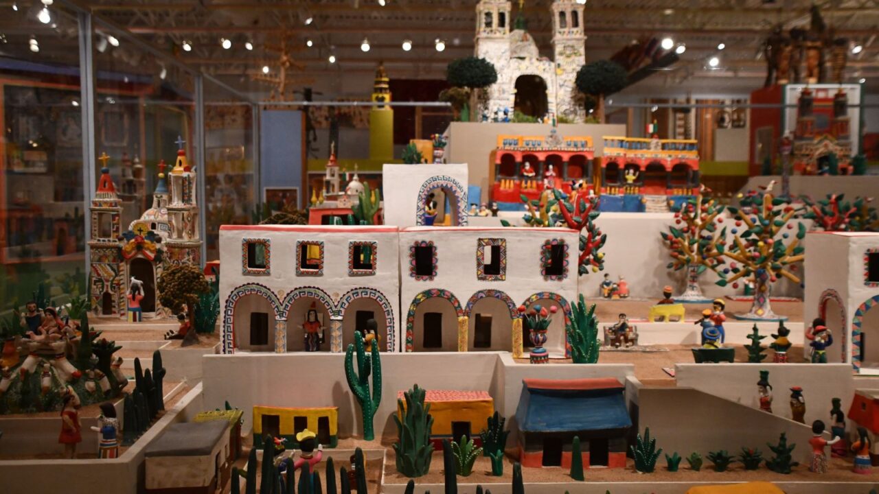 A detailed miniature village scene display, featuring numerous small buildings, figures, and elements like cacti and trees, under bright lighting in what appears to be a museum exhibit.