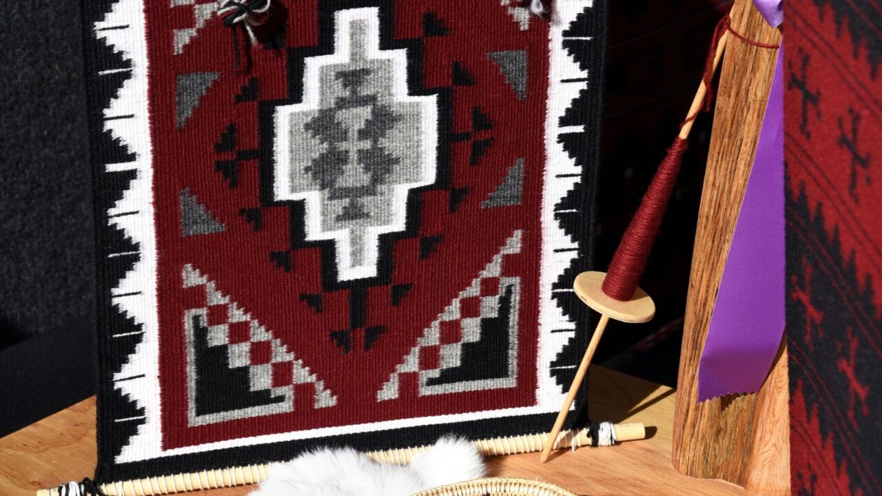 A traditional Navajo loom with a partially completed red, white, and black geometric patterned rug, alongside a basket of yarn and a white sheepskin rug on a wooden surface.