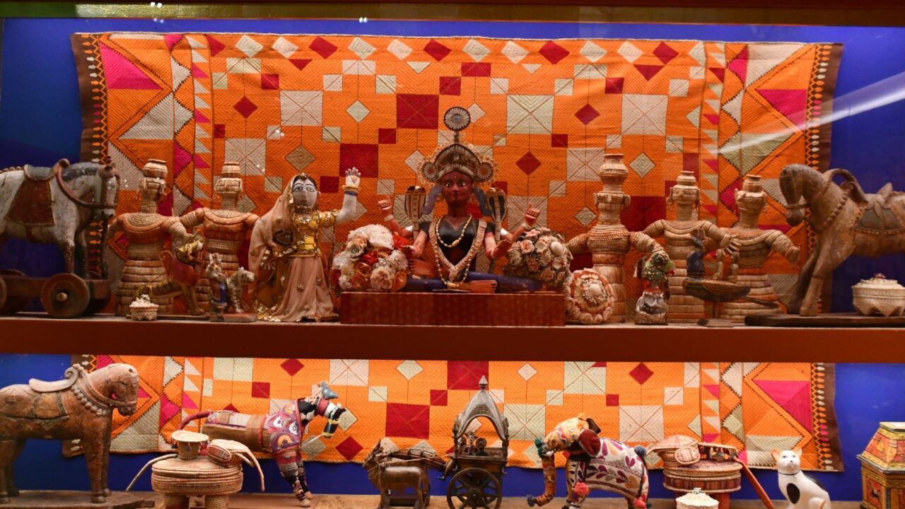 A colorful display of folk art artifacts, including statues of deities and figures, animal sculptures like horses and camels, and decorative pots, arranged on wooden shelves in front of a vibrant, patterned textile wall hanging.