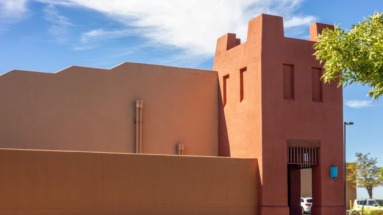 An adobe-style building with a warm, reddish-brown stucco exterior under a bright blue sky with white clouds.