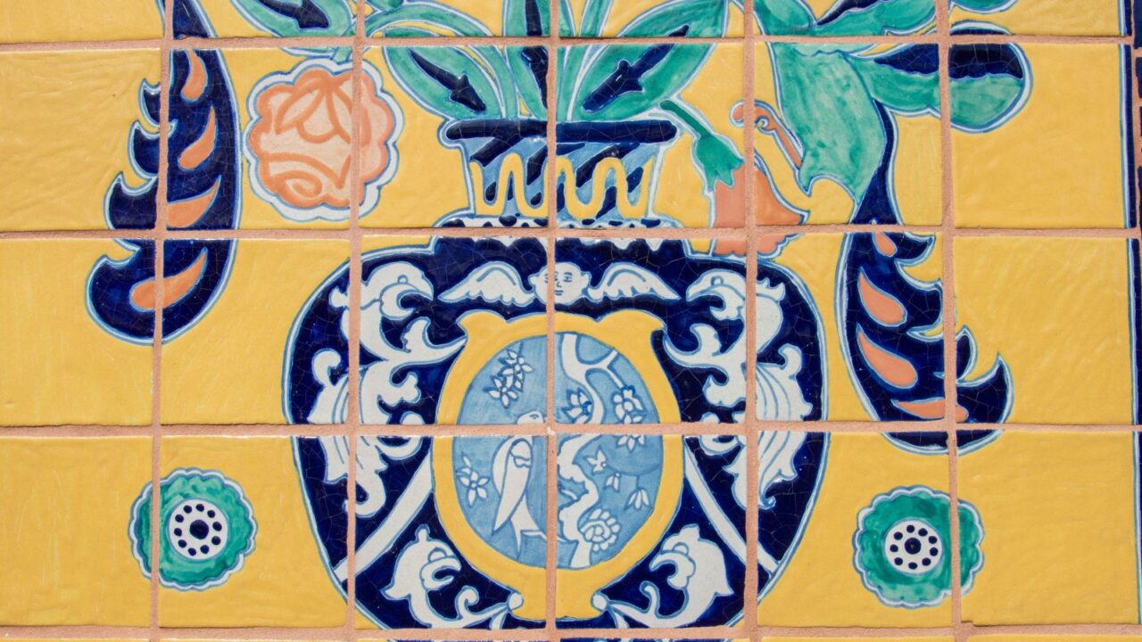 A close-up of colorful Talavera Poblana tiles, featuring a central vase design with elaborate blue and white patterns, surrounded by yellow tiles with orange and green floral motifs.