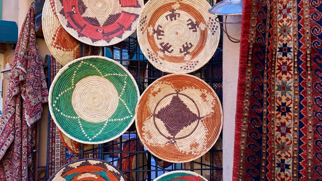 Colorful woven baskets with traditional Native American patterns, created by Apache, Hopi, and Tohono O’odham artists, are displayed on a black metal grid at a Santa Fe museum, surrounded by patterned textiles and rugs.