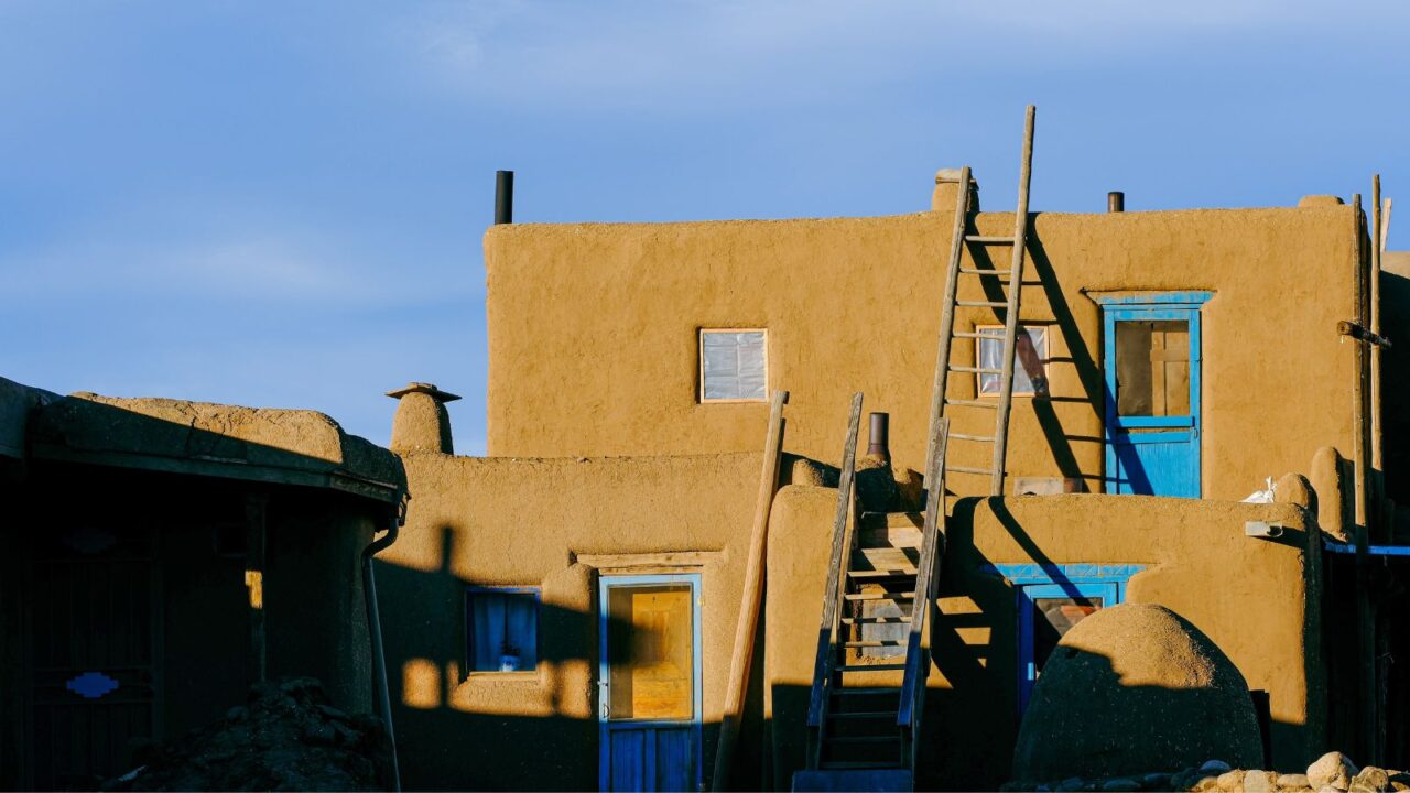 An adobe building with brown walls and prominent blue doors and windows, featuring wooden ladders leaning against its multi-story structure, under a clear sky.
