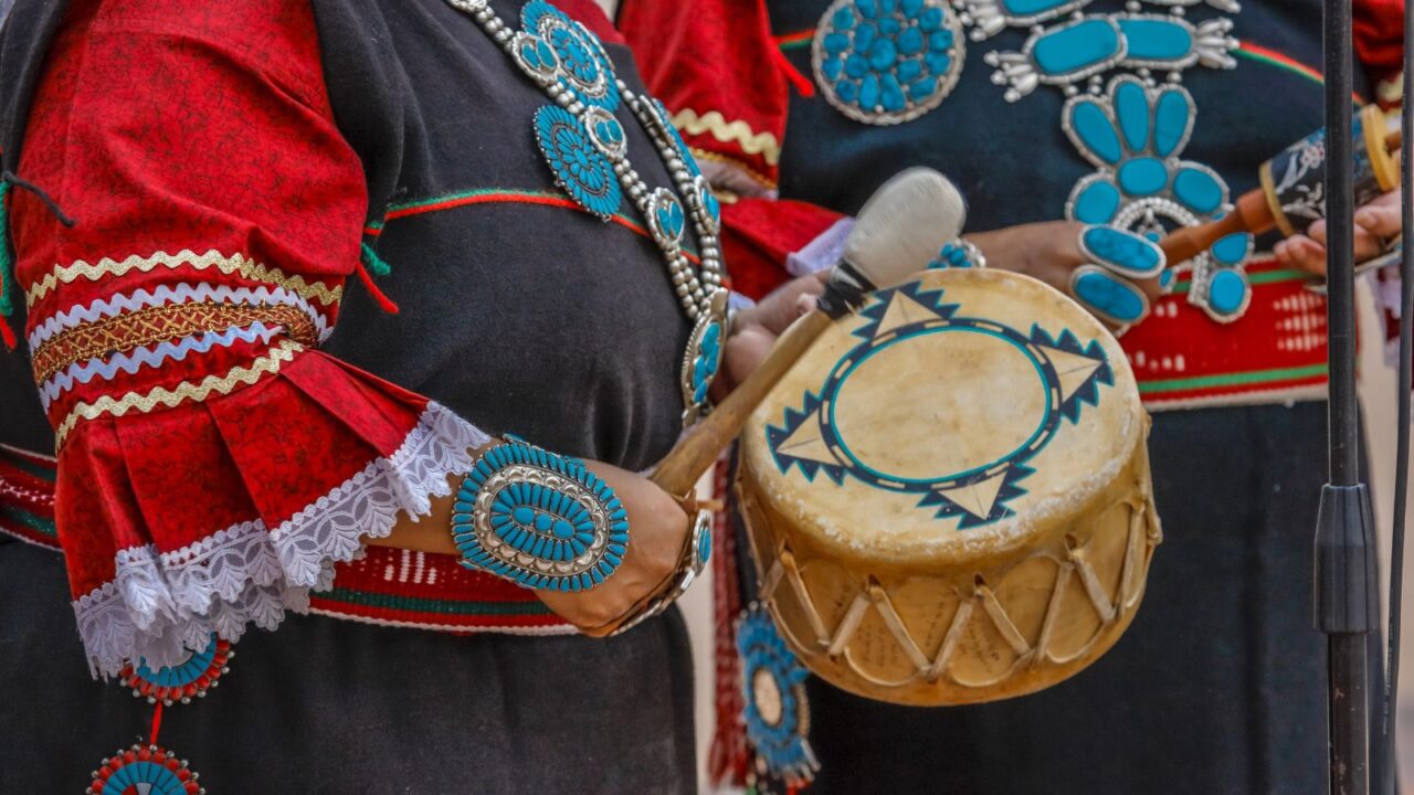 Close-up of a person in traditional Zuni attire, featuring turquoise and silver jewelry, holding a drum and drumstick during a ceremony in New Mexico.