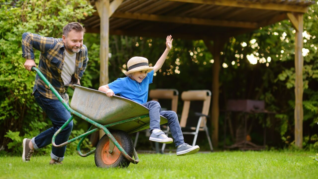 A man pushing a smiling child in a wheelbarrow across a grassy yard with a wooden shelter in the background.
