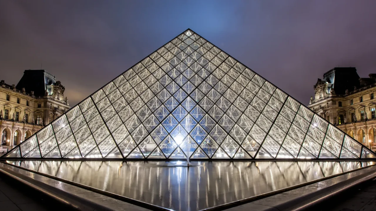 Night view of the illuminated glass pyramid entrance of the Louvre Museum in Paris, reflecting in the surrounding water with historic buildings in the background.
