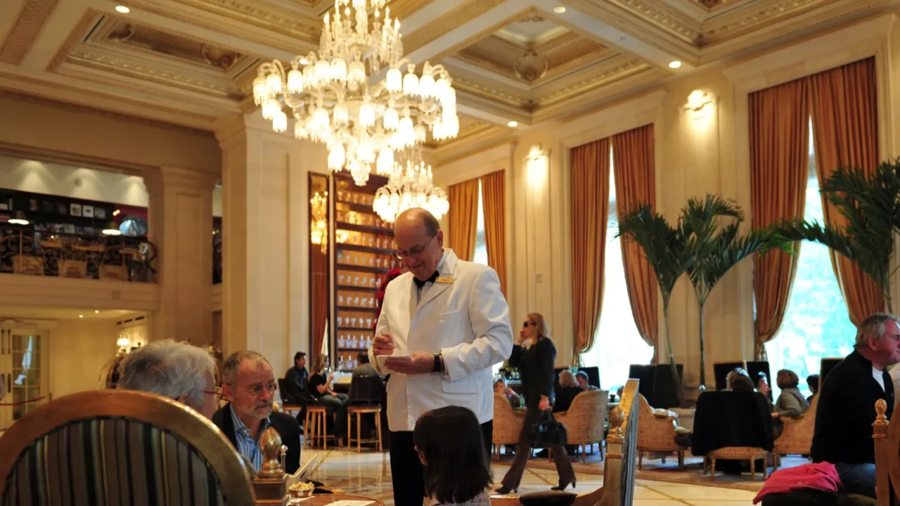 Restaurant in the Plaza Hotel featuring crystal chandelier, wall sconces, floor to ceiling windows with curtains, filled with people enjoying their meals and servers.