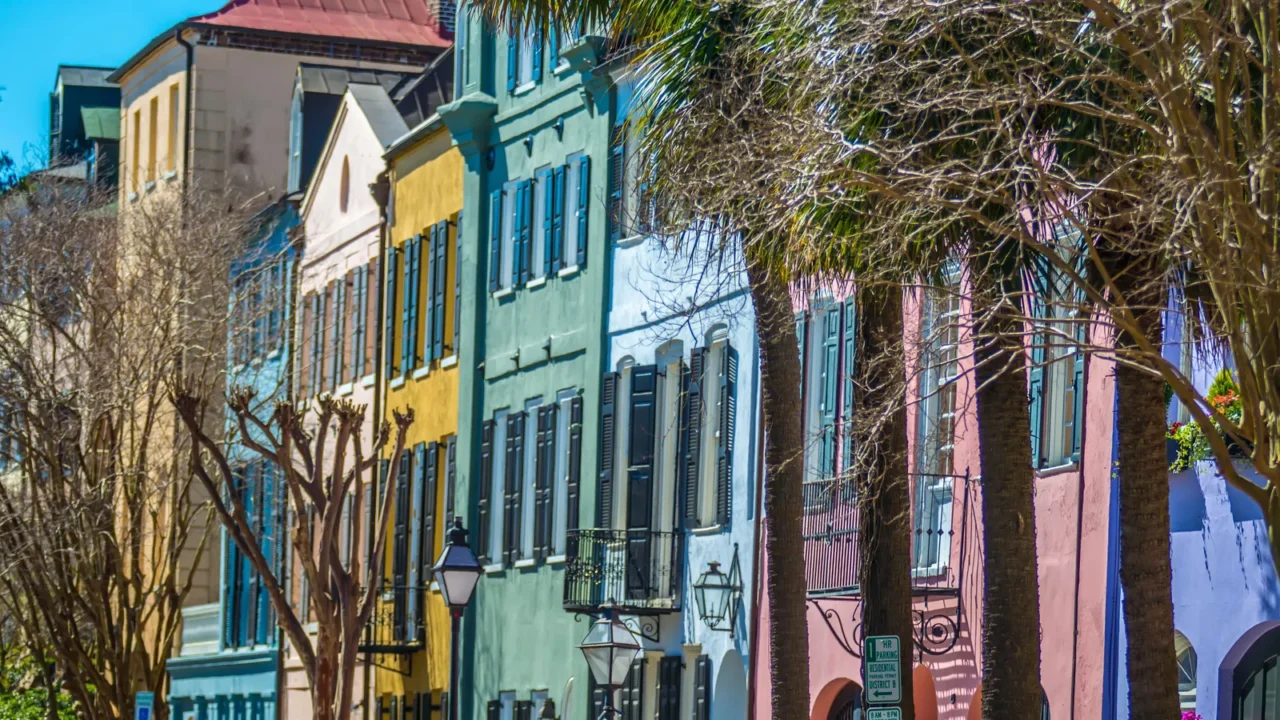 A row of historic, colorful houses known as Rainbow Row in Charleston, South Carolina, with palm trees and bare trees in the foreground.
