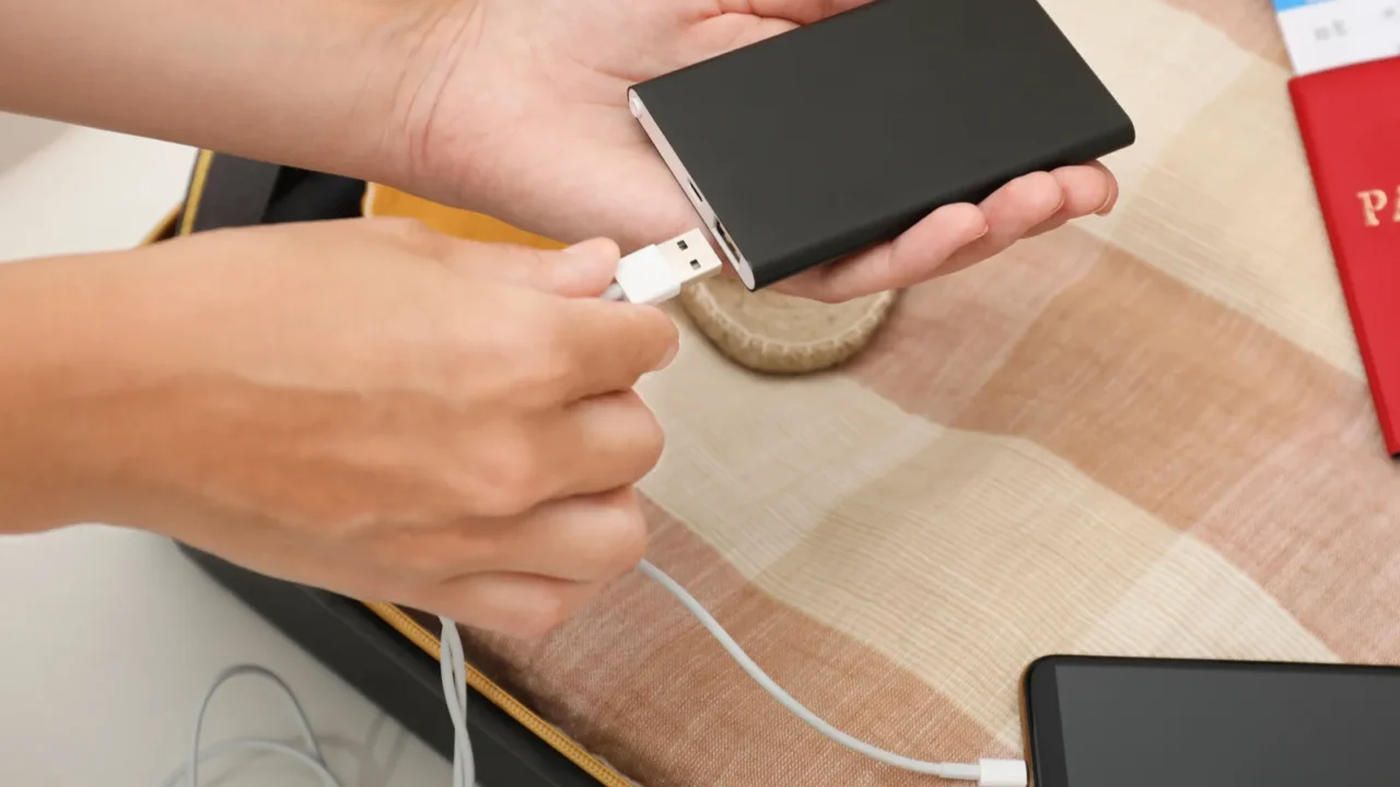 woman charging smartphone with power bank near packed suitcase indoors