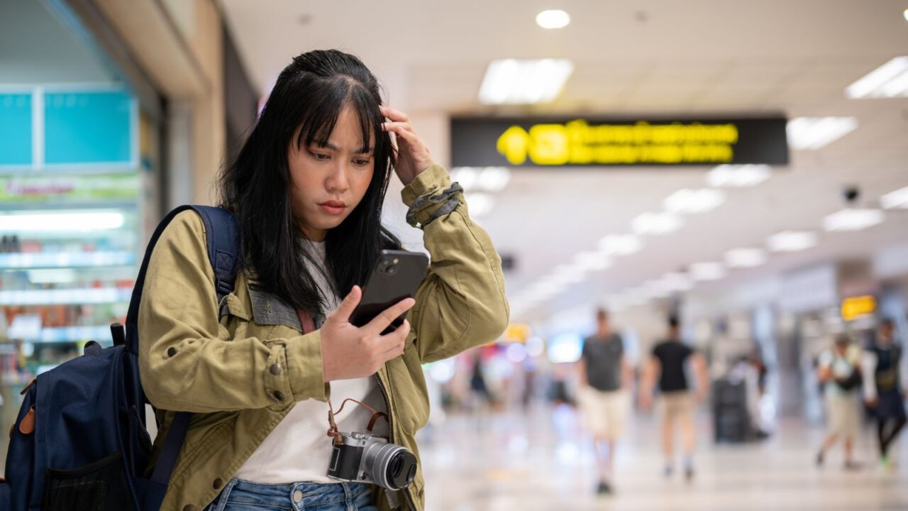 A confused young Asian female solo traveler is reading her flight information on her phone with a serious, doubtful face, facing a problem with self-check-in or missing