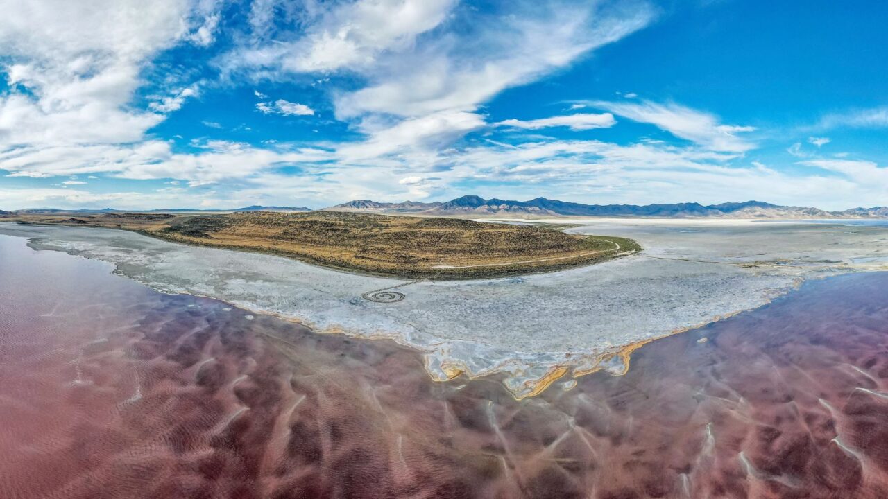 Great Salt Lake, pinkish water, a land mass called the Promontory Mountains, an artwork called the Spiral Jetty. In the distance, mountains and clouds.