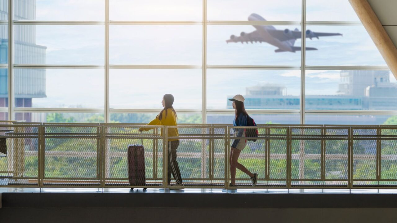Two happy Asian girls traveling abroad together, carrying suitcase luggage and running through big window of airport.