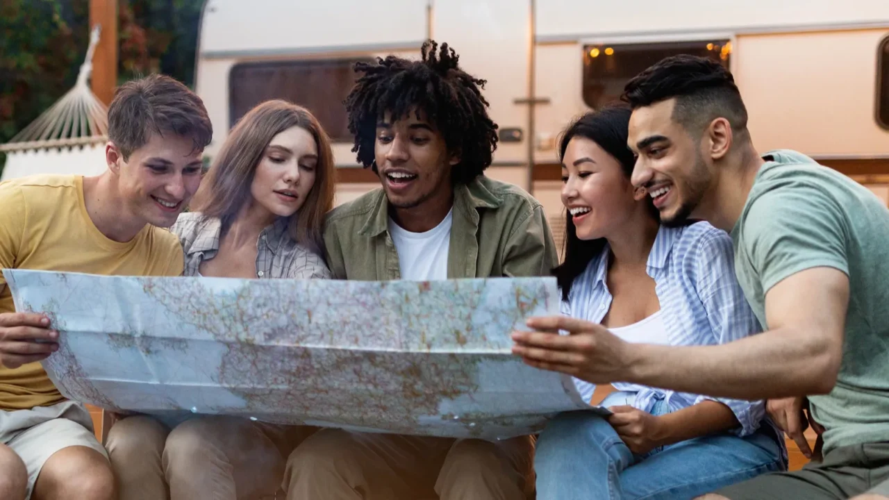 happy multiracial young people sitting near campfire checking map finding