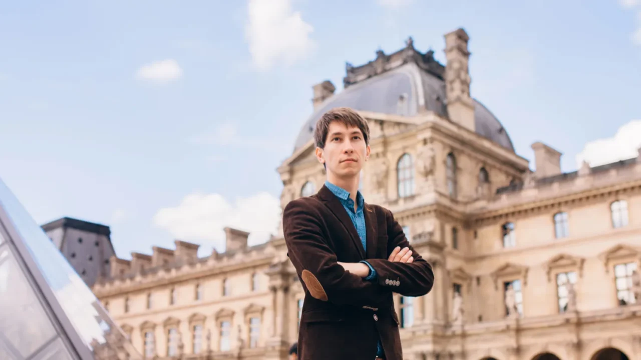 a young man stands against the background of the louvre