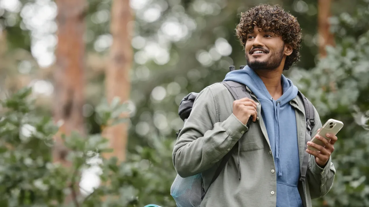carefree young indian hiker with backpack holding smartphone and looking