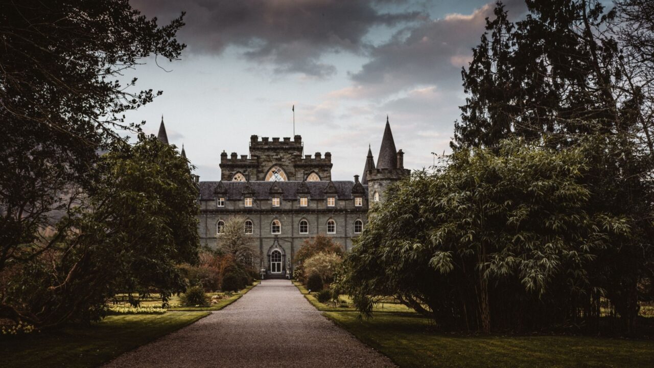 The image shows a Scottish castle surrounded by forest, with impressive stone walls and tall towers. A beautiful entrance with stone steps leads to the main building, creating a majestic impression.