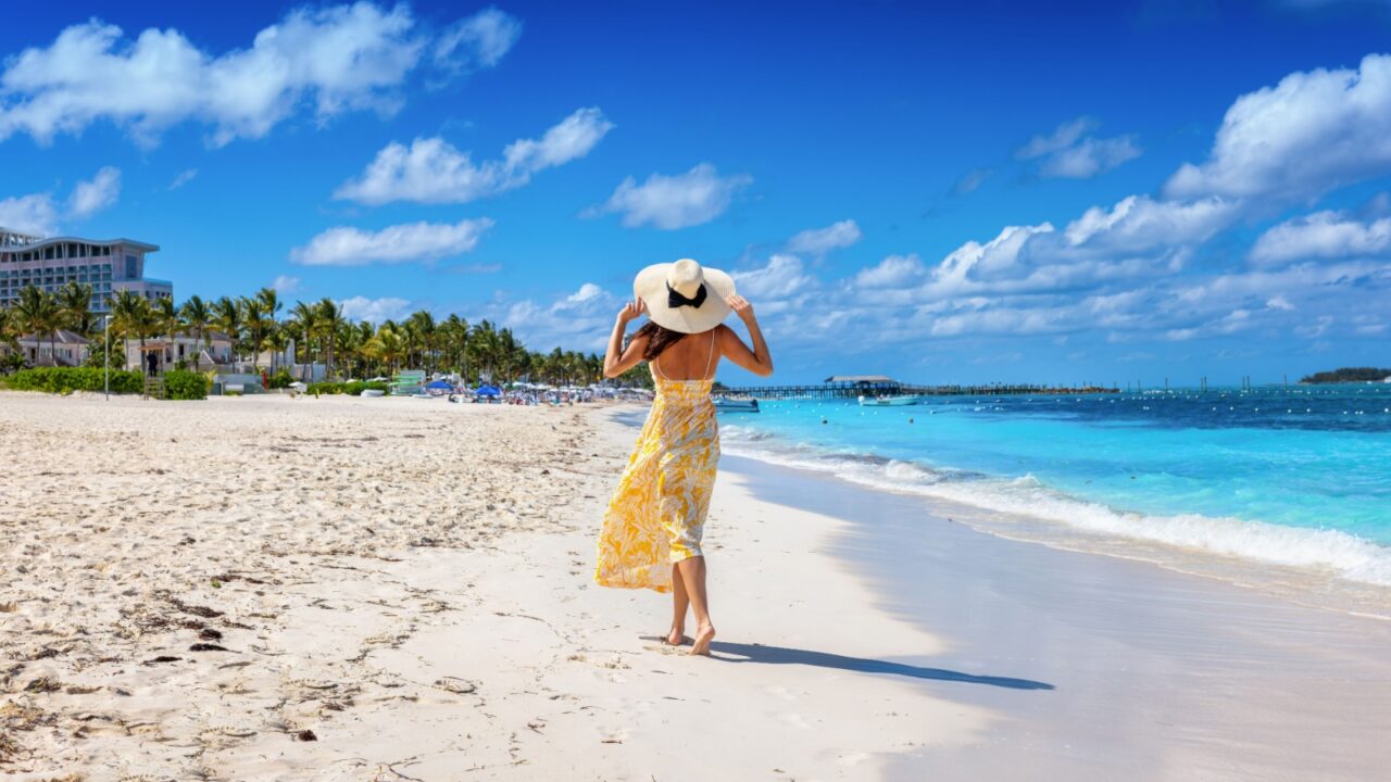 A happy holiday woman in a yellow summer dress walks down a tropical beach at Goodman's Bay in Nassau, Bahamas, The Caribbean Sea