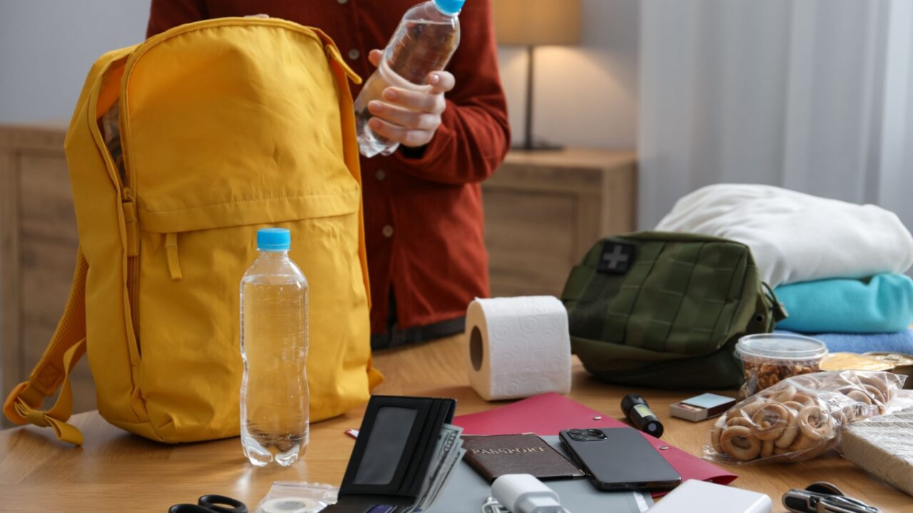 Survival kit. Woman packing different emergency supplies into backpack at wooden table indoors, closeup
