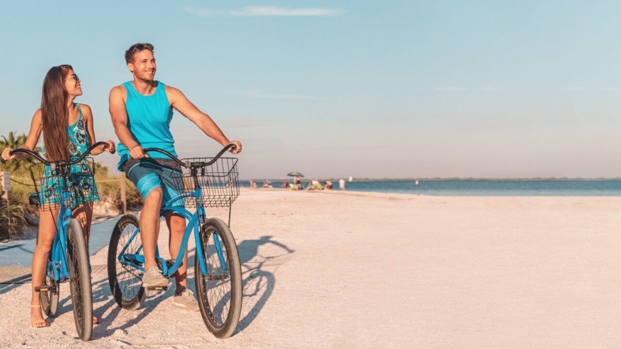 Florida beach vacation couple biking sport rental bikes recreational activity happy watching sunset on Sanibel Island by the Lighthouse. Young woman and man riding bicycles. Summer people lifestyle.