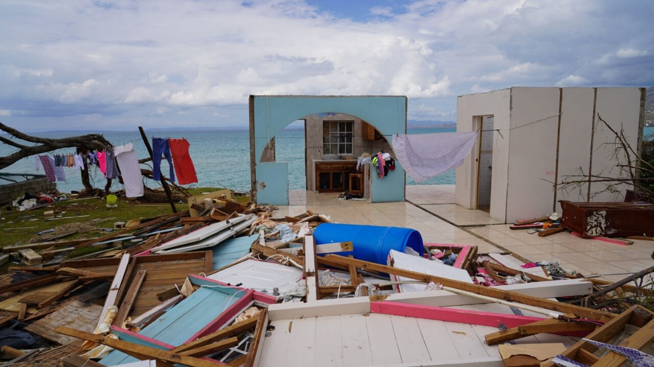 The aftermath of Hurricane Melissa is depicted, showing a house ripped apart with debris scattered across the landscape, while the remnants of a life, like laundry on a line, cling to the seaside.