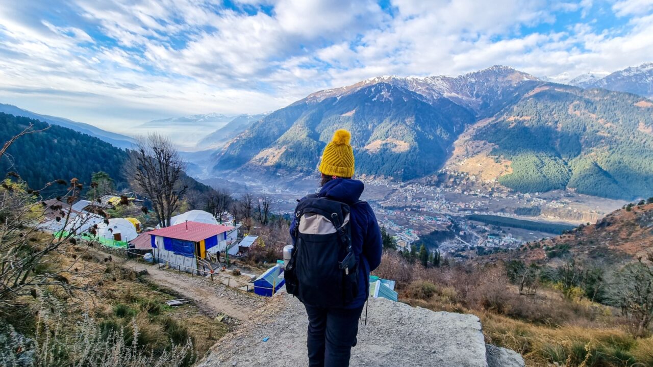 Female trekker hiker standing looking down at village houses and himalaya mountains in distance and cloudy sky in manali, kullu, shimla, kedarnath in India