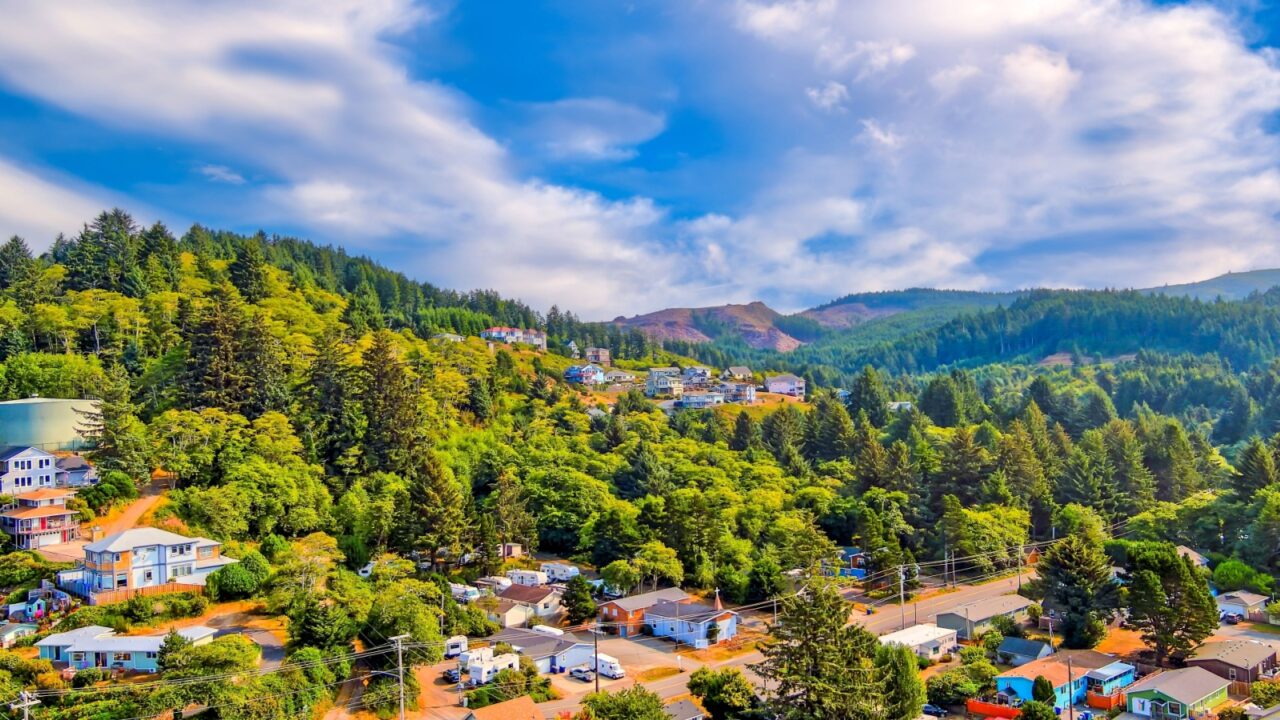 Aerial view of Depoe Bay, Oregon with rolling hills with beautiful houses nestled amongst tall trees in late summer with a partly cloudy blue sky.