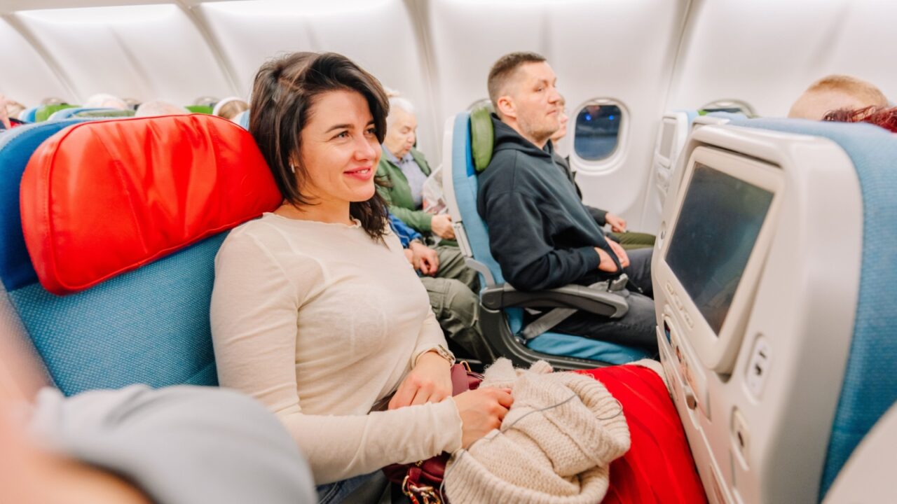 Riga, Latvia - February 22, 2025 - A smiling woman sits comfortably in an airplane seat with a red headrest, holding a blanket. Other passengers are seated, and screens are visible.
