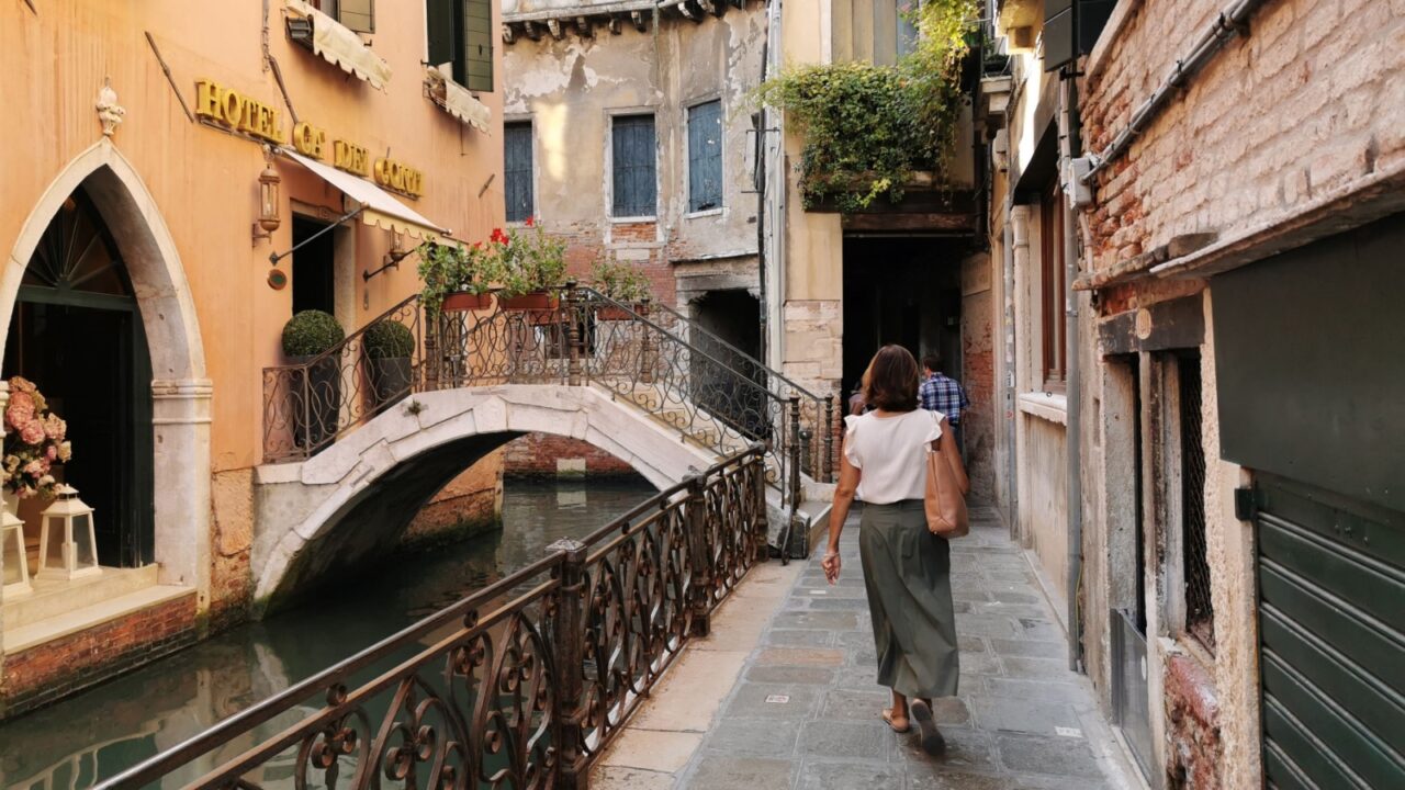 Venice, Veneto, Italy - September 11, 2023: A woman walks along a narrow cobblestone alley beside a canal, passing historic Venetian buildings with aged facades and shuttered windows