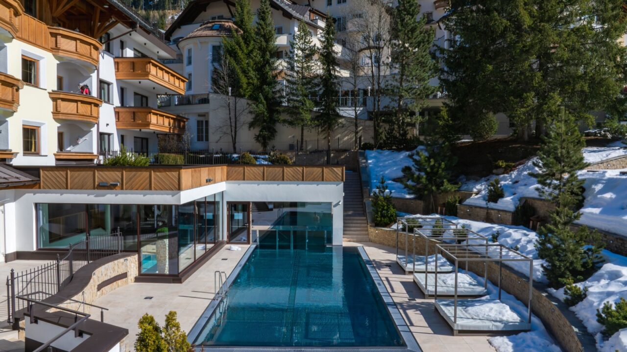 Modern outdoor pool with lounge chairs at a luxury hotel in Ischgl, Austria, surrounded by snow, evergreen trees, and distant ski lift structures.